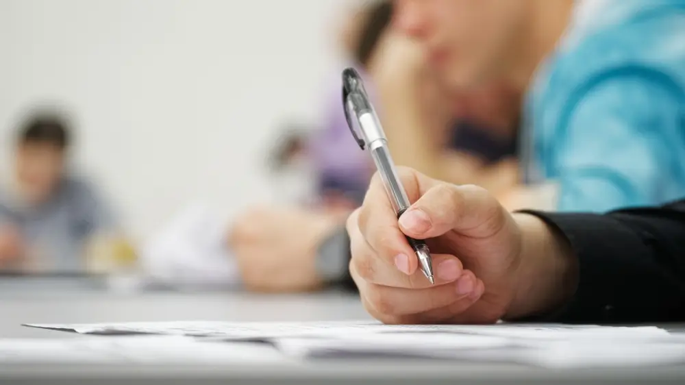 Primo piano di una mano che scrive con una penna durante la prova scritta di un concorso pubblico in aula.