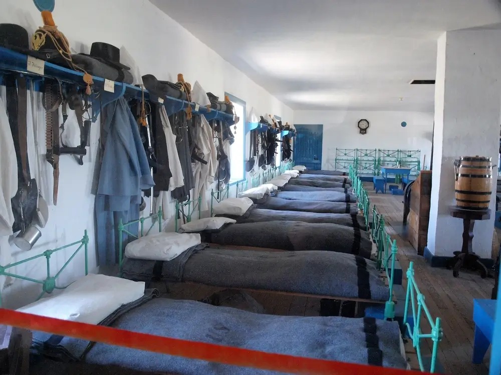Camerata di una caserma militare con letti in fila e uniformi appese, simile a un centro di selezione dell'Esercito.